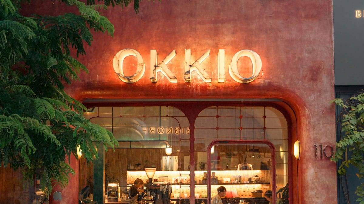越南一家咖啡店的夜間門面，OKKIO 霓虹招牌亮著暖光，玻璃窗內有店員與顧客