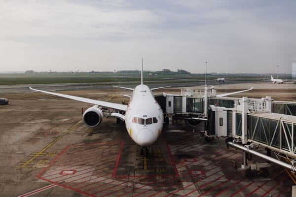 機場停機坪：一架白色客機正面停靠空橋，地勤在機翼下作業，遠處還有飛機在滑行