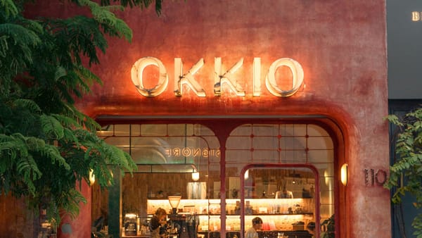 越南一家咖啡店的夜間門面，OKKIO 霓虹招牌亮著暖光，玻璃窗內有店員與顧客
